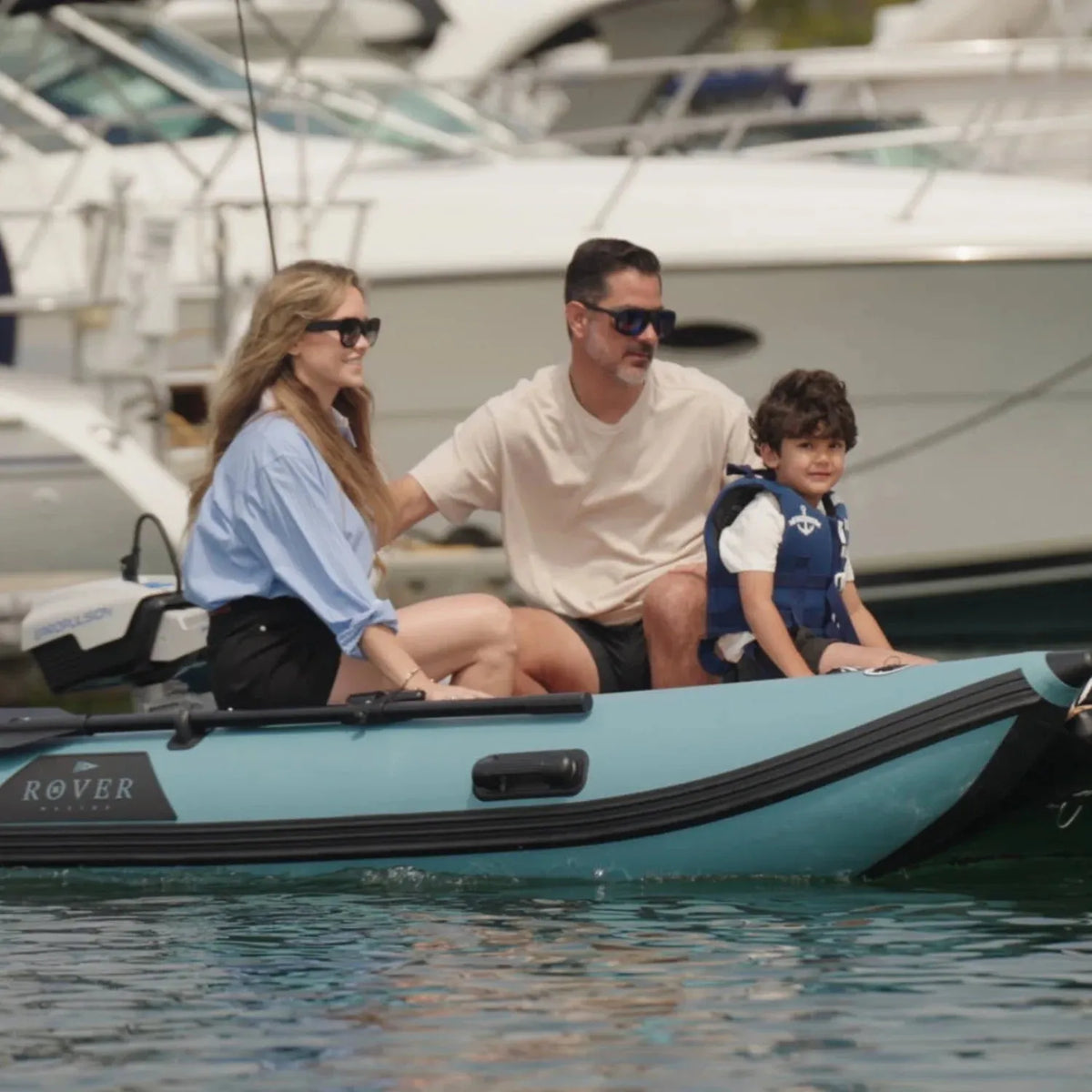 Family of three on a small boat in a marina with yachts in the background
