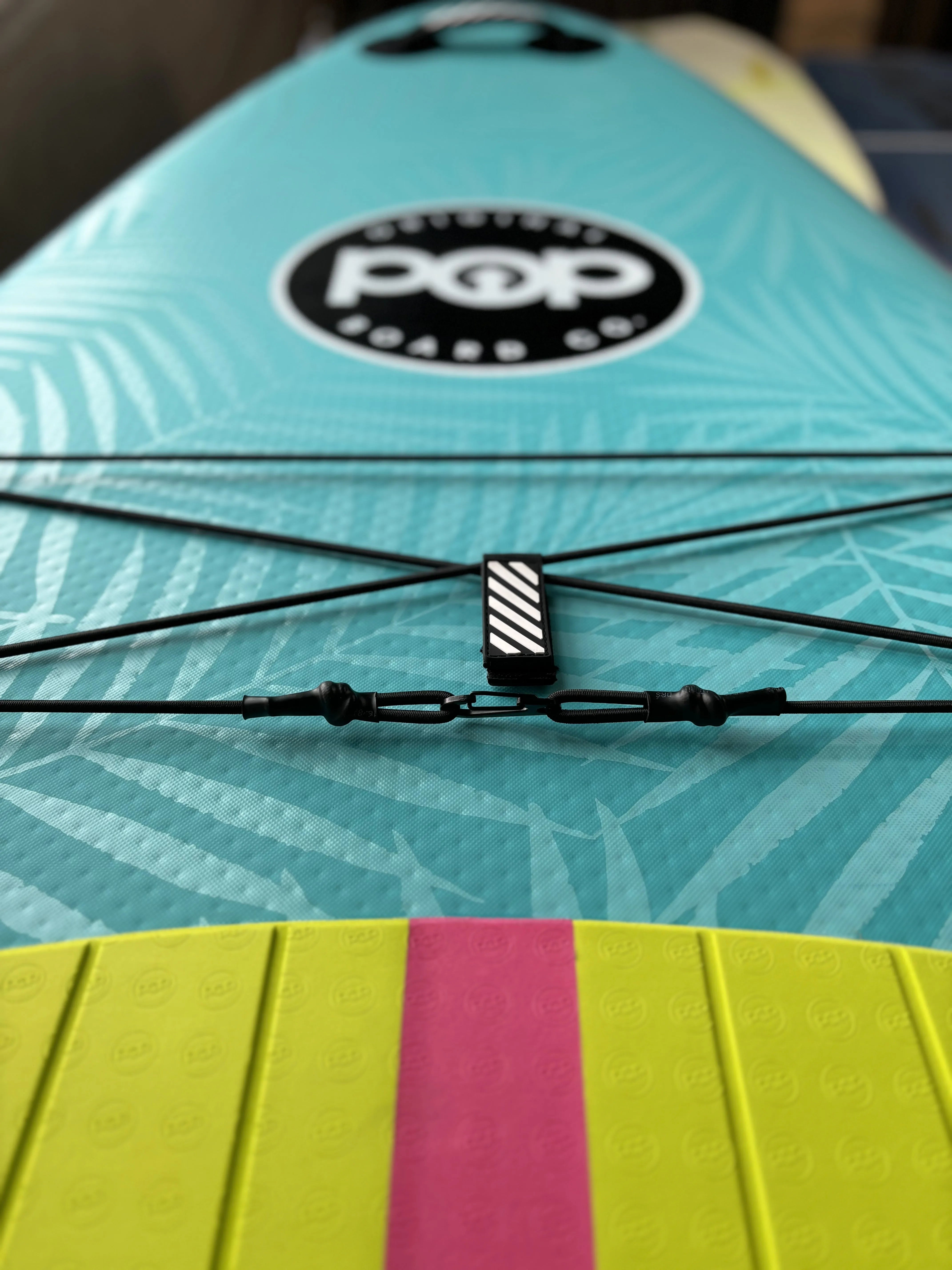Close-up of the front cargo bungees on the turquoise Yacht Hopper paddleboard, showing the black elastic cords, palm leaf print deck, and POP Board Co. logo.